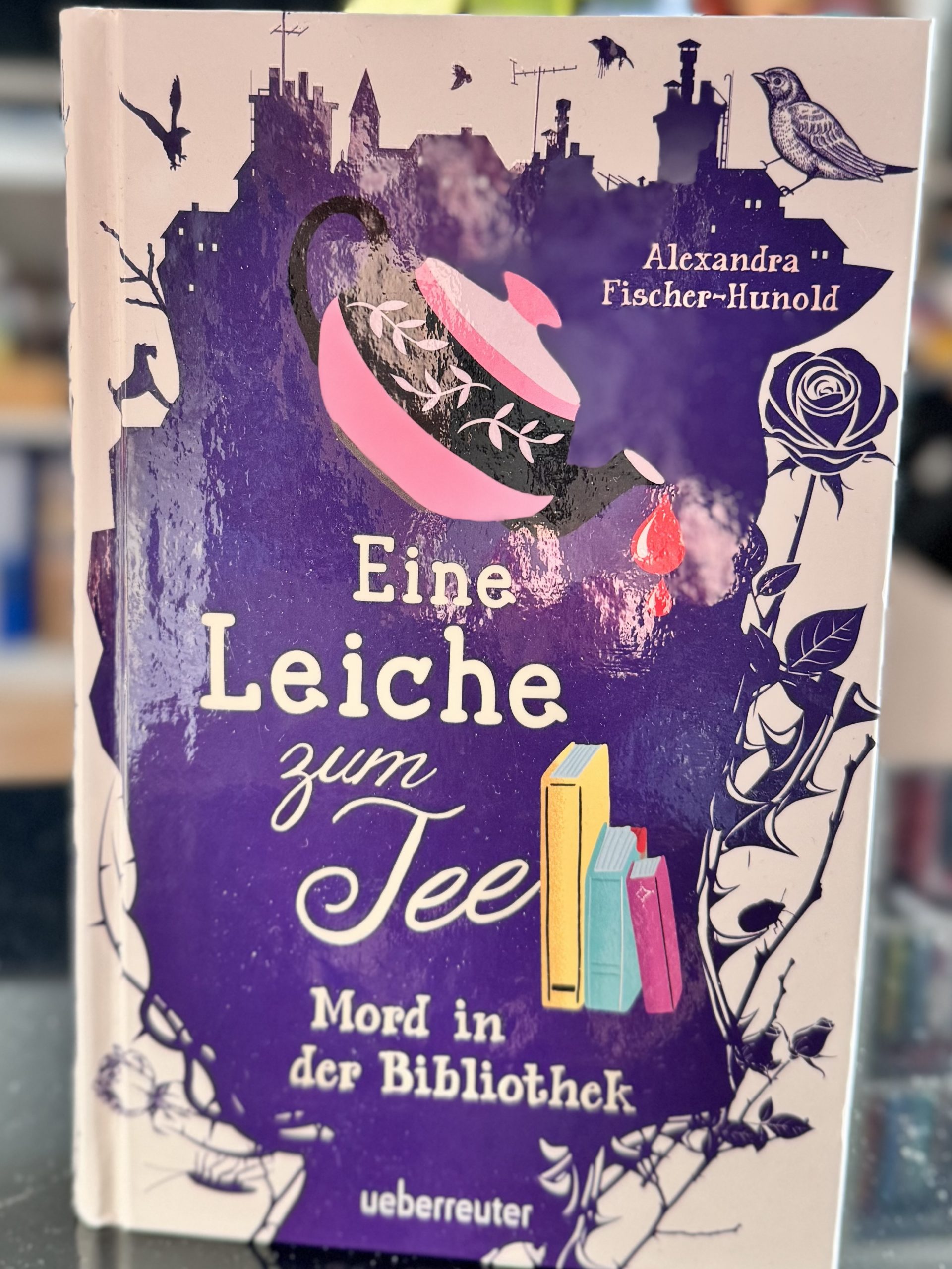 Auf dem Cover schüttet eine rosafarbene Teekanne Bluttropfen auf drei Bücher.
