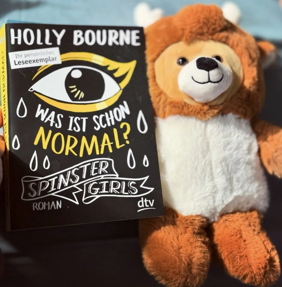 Ein Stoffbär der das Buch "Was ist schon normal" festhält.