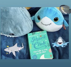 Mit dir am Meer / Shark Club 1 Das türkise Cover mit blauem Titel. Daneben liegen zwei Stofftier-Haie.