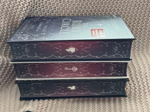 Der Buchschnitt der Ruby Circle Trilogie in grau-blau und rot-lila Tönen verziert mit geschwungenen weißen Linien.