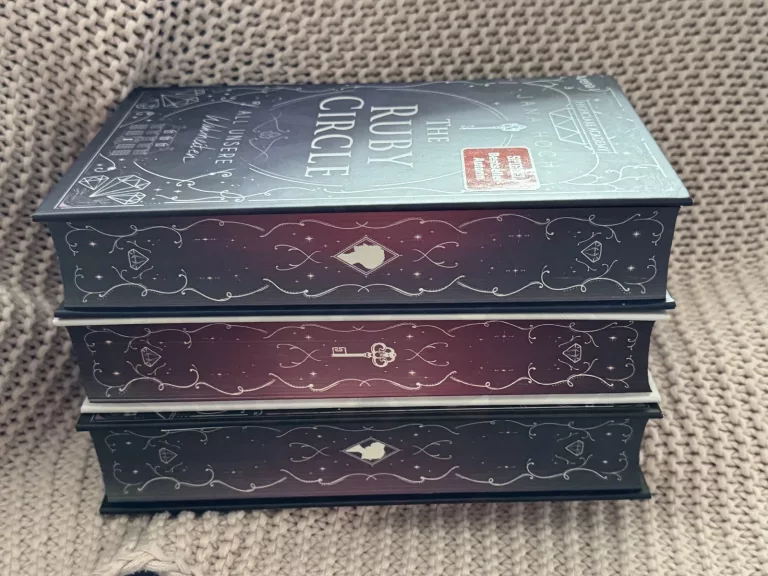 Der Buchschnitt der Ruby Circle Trilogie in grau-blau und rot-lila Tönen verziert mit geschwungenen weißen Linien.
