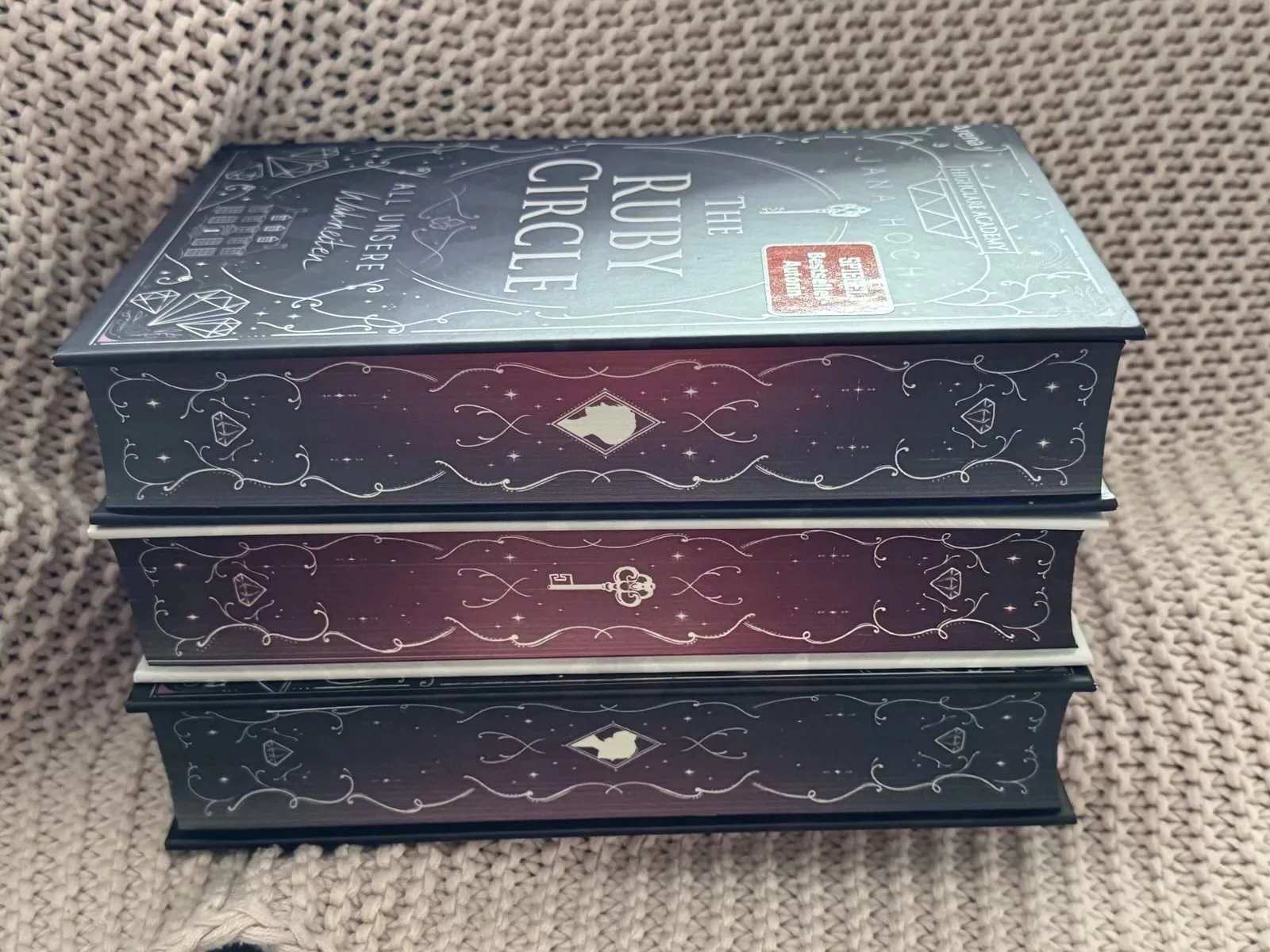 Der Buchschnitt der Ruby Circle Trilogie in grau-blau und rot-lila Tönen verziert mit geschwungenen weißen Linien.