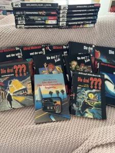 Viele verschiedene "Die drei ???" Bücher liegen verstreut auf einer Couch.