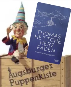 Herzfaden 1 „Buch ‚Herzfaden‘ von Thomas Hettche über die Augsburger Puppenkiste mit Kasperlfigur die uf einer Holzkiste mit dem Logo der berühmten Marionettenbühne sitzt.“