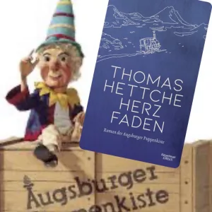 „Buch ‚Herzfaden‘ von Thomas Hettche über die Augsburger Puppenkiste mit Kasperlfigur die uf einer Holzkiste mit dem Logo der berühmten Marionettenbühne sitzt.“