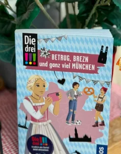 Auf dem hellblauen Cover sind die drei Ausrufezeichen mit Brezen und Dirndl oder Lederhose zu sehen.
