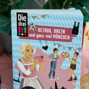 Auf dem hellblauen Cover sind die drei Ausrufezeichen mit Brezen und Dirndl oder Lederhose zu sehen.