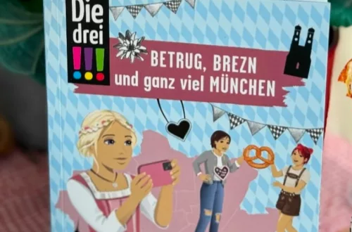 Auf dem hellblauen Cover sind die drei Ausrufezeichen mit Brezen und Dirndl oder Lederhose zu sehen.