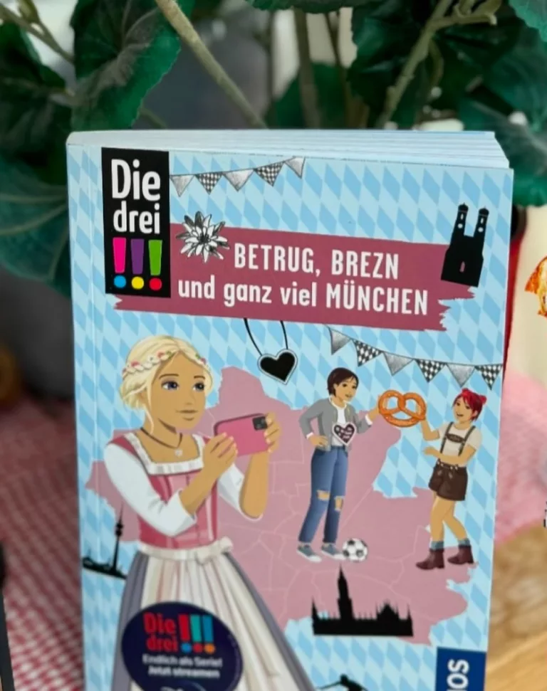 Auf dem hellblauen Cover sind die drei Ausrufezeichen mit Brezen und Dirndl oder Lederhose zu sehen.