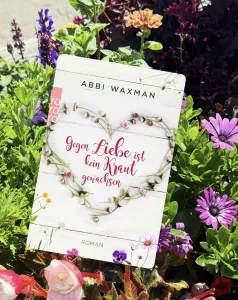 Buchcover des Romans „Gegen Liebe ist kein Kraut gewachsen“ von Abbi Waxman, fotografiert in einem Blumenbeet mit bunten Blüten. Das Cover zeigt einen aus Blüten geformten Herzkranz auf weißem Holz.