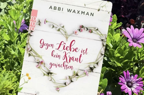 Buchcover des Romans „Gegen Liebe ist kein Kraut gewachsen“ von Abbi Waxman, fotografiert in einem Blumenbeet mit bunten Blüten. Das Cover zeigt einen aus Blüten geformten Herzkranz auf weißem Holz.