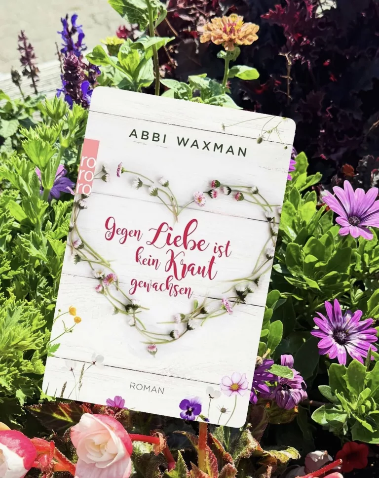 Buchcover des Romans „Gegen Liebe ist kein Kraut gewachsen“ von Abbi Waxman, fotografiert in einem Blumenbeet mit bunten Blüten. Das Cover zeigt einen aus Blüten geformten Herzkranz auf weißem Holz.