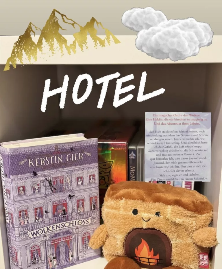 „Ein herbstlich gestaltetes Bücherregal mit dem Roman ‚Wolkenschloss‘ von Kerstin Gier im Vordergrund. Neben dem Buch sitzt ein braunes Plüschtier in Form eines lodernden Kaminfeuers mit einem freundlichen Gesicht. Im Hintergrund ist ein Schild mit der Aufschrift ‚HOTEL‘, eine Textpassage aus dem Buch sowie Deko-Elemente wie goldene Berge und eine Wolke zu sehen.“