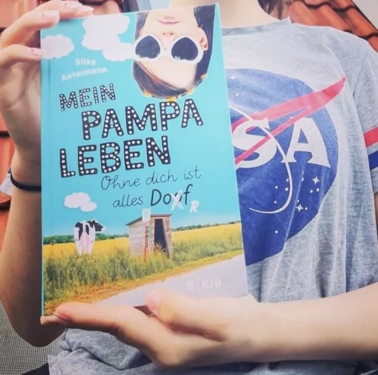 Mein Pampa Leben 1 Eine Person hält das Jugendbuch „Mein Pampaleben – Ohne dich ist alles Dorf“ von Silke Antelmann in den Händen. Das Cover ist hellblau und zeigt ein Mädchen mit Sonnenbrille, das kopfüber ins Bild ragt, sowie eine Kuh auf einer Wiese im Hintergrund. Die Person trägt ein graues T-Shirt mit dem NASA-Logo.