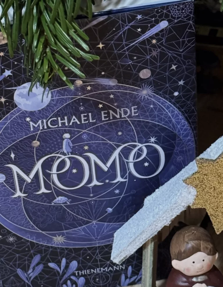 Das Cover des Buches „Momo“ von Michael Ende in einer dunkelblauen, astronomischen Gestaltung. Im Zentrum steht der große, geschwungene Schriftzug „MOMO“. Darüber ist eine kleine, hell gezeichnete Figur von oben zu sehen, die auf einer kreisförmigen Umlaufbahn balanciert. Eine kleine Schildkröte kriecht im unteren Bereich des Kreises. Der Hintergrund zeigt ein komplexes Muster aus Sternenbildern, Planetenbahnen, Sternen und einer Sternschnuppe. Am oberen linken Rand ragen grüne Tannenzweige ins Bild, und unten rechts ist die Spitze eines glitzernden Holzsterns sowie der Kopf einer kleinen Deko-Figur zu erkennen. Der Verlagsname „THIENEMANN“ steht ganz unten in weißer Schrift.
